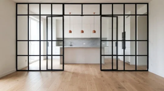 Intérieur contemporain lumineux avec verrière d'atelier séparant cuisine blanche et espace de vie, parquet chêne clair et suspensions cuivre